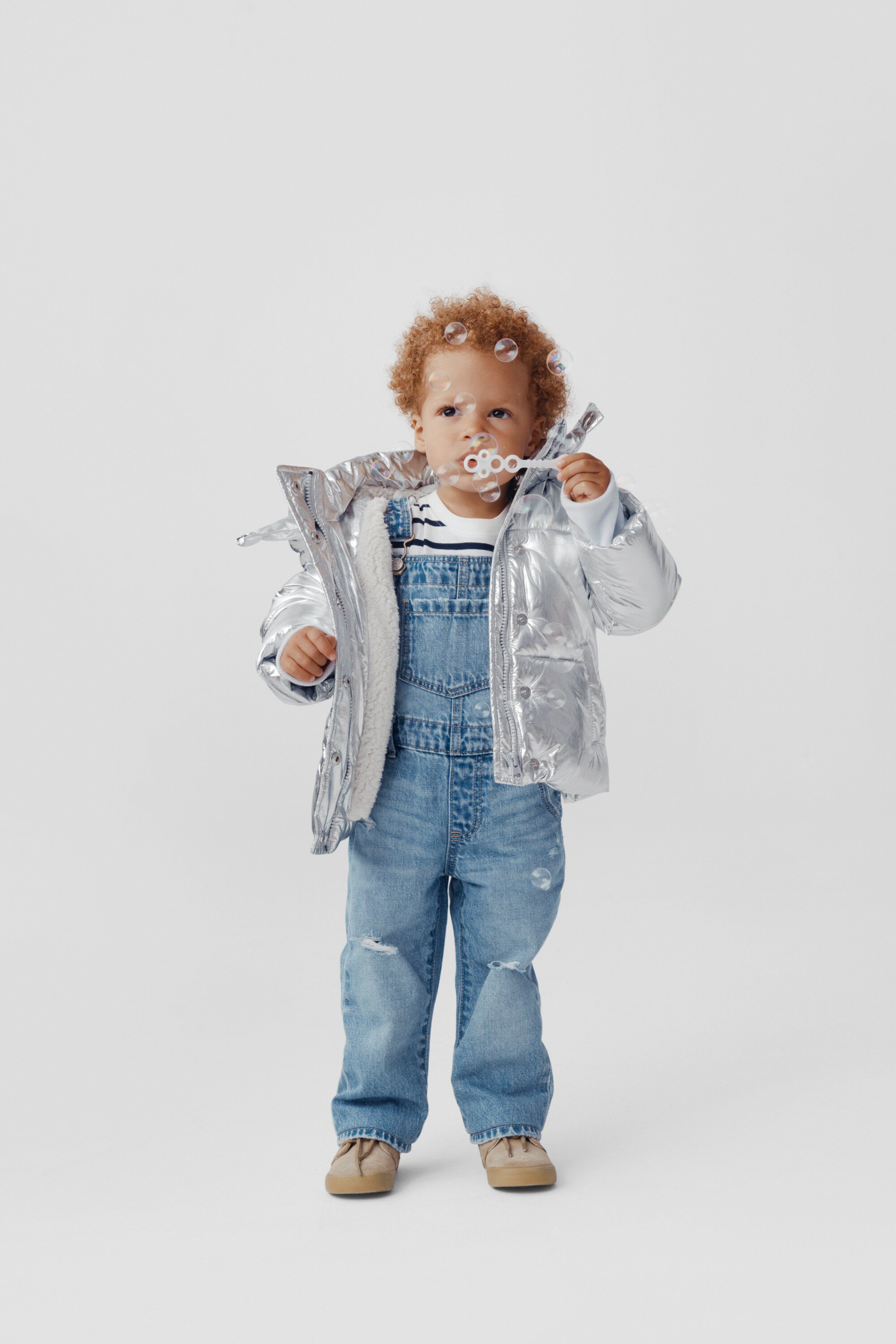 A full shot of a toddler with curly red hair blowing bubbles while wearing a metallic silver puffer jacket and denim overalls on white.