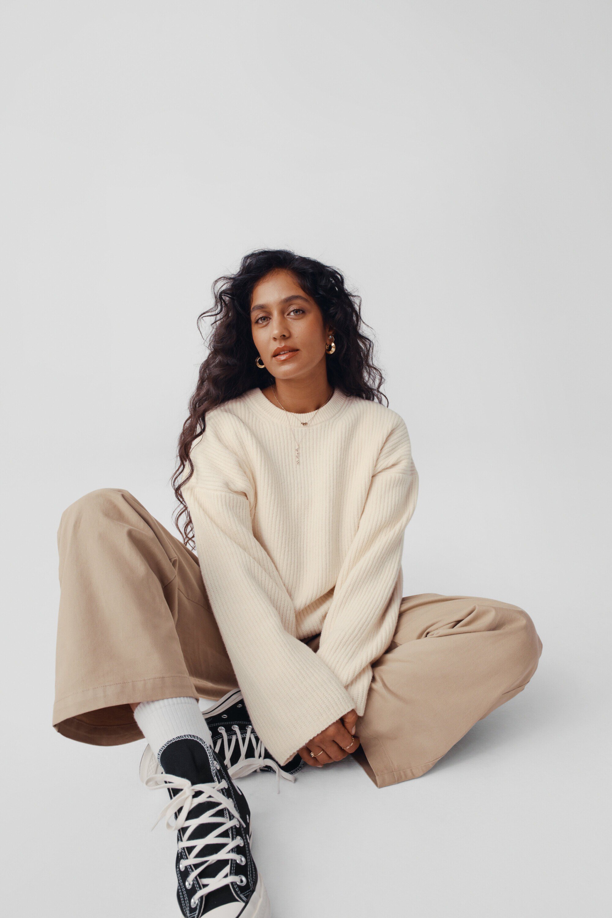 Adult woman with long dark curly hair sits cross-legged in a photo studio wearing a cream ribbed sweater, khaki pants, and black Converse sneakers.