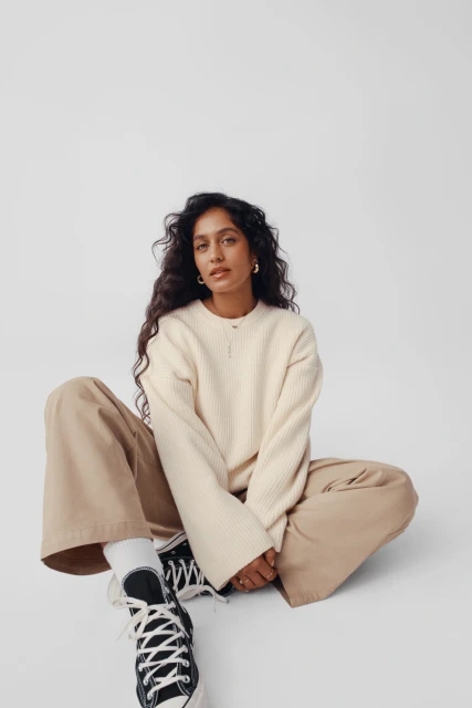 Adult woman with long dark curly hair sits cross-legged in a photo studio wearing a cream ribbed sweater, khaki pants, and black Converse sneakers.