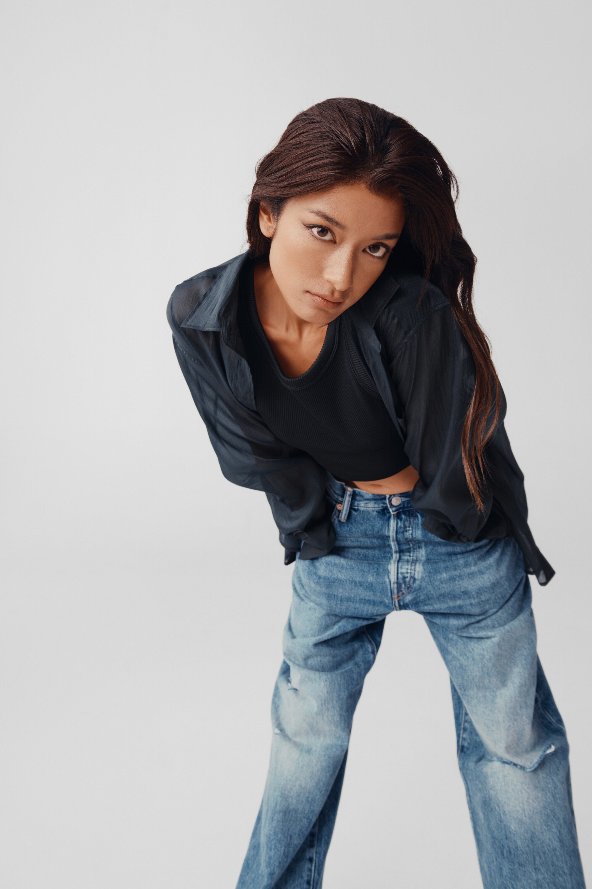 A woman with long brown hair poses indoors wearing a black t-shirt, blue denim jeans, and a black button-down shirt.