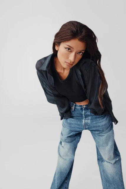 A woman with long brown hair poses indoors wearing a black t-shirt, blue denim jeans, and a black button-down shirt.