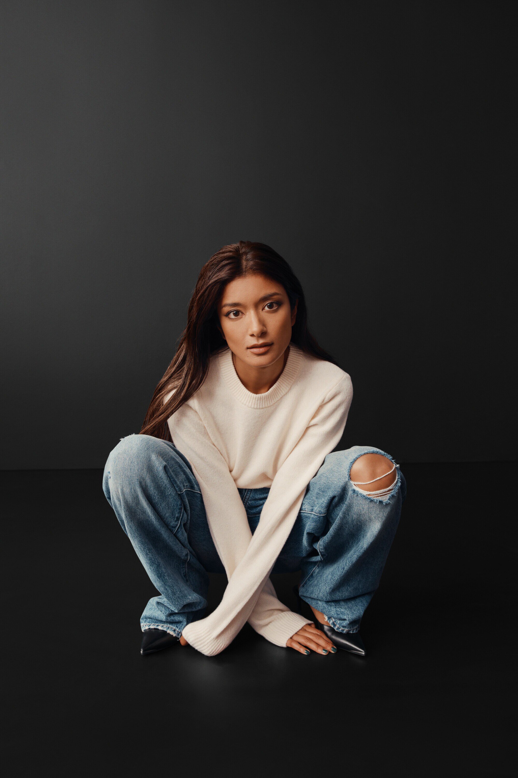 Portrait of a young woman with long brown hair, wearing a cream sweater and ripped jeans, crouching on a dark gray background.