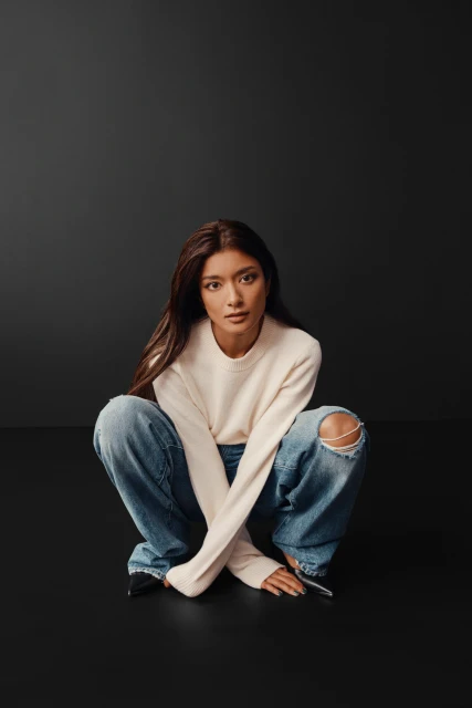 Portrait of a young woman with long brown hair, wearing a cream sweater and ripped jeans, crouching on a dark gray background.