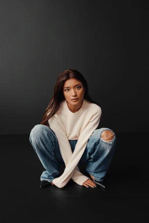 Portrait of a young woman with long brown hair, wearing a cream sweater and ripped jeans, crouching on a dark gray background.