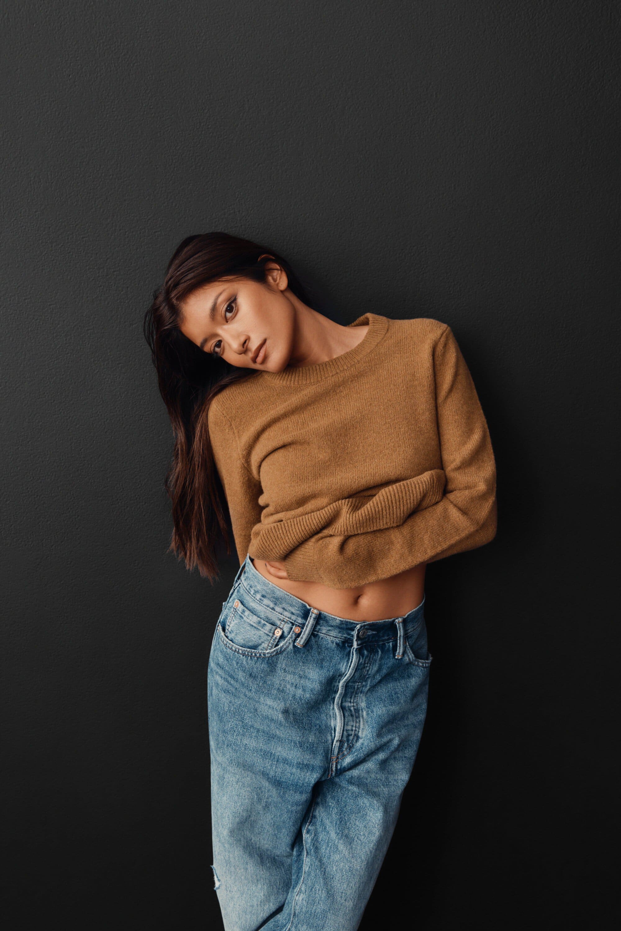 Brunette woman in a caramel sweater and denim jeans leans against a charcoal gray wall in a medium studio shot for fashion.