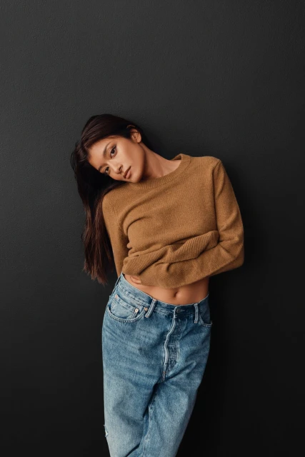 Brunette woman in a caramel sweater and denim jeans leans against a charcoal gray wall in a medium studio shot for fashion.