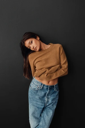 Brunette woman in a caramel sweater and denim jeans leans against a charcoal gray wall in a medium studio shot for fashion.