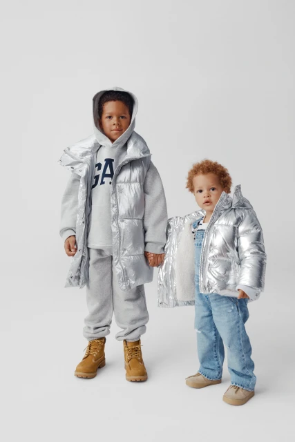 Two children, one with darker skin in silver puffer vest and grey sweat suit, the other in a silver puffer jacket and overalls.