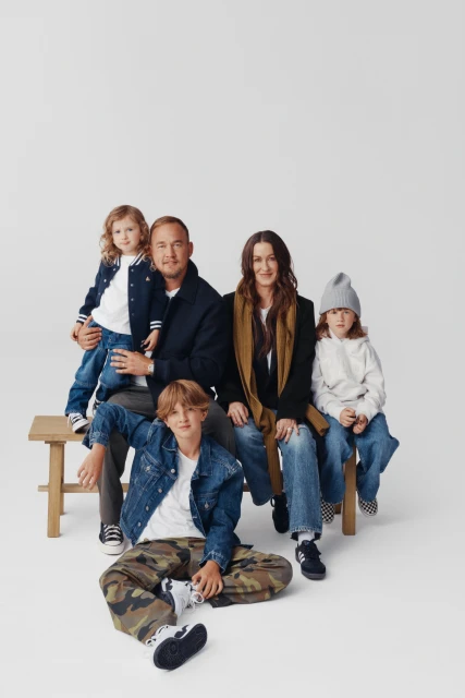 A family of five poses for a portrait in a bright studio, wearing denim and neutral tones, seated and standing on a wooden bench.