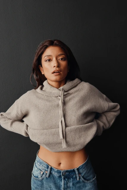 Attractive Asian woman in a cropped hooded sweater poses in front of a dark wall, her hands on her hips and her jeans visible.
