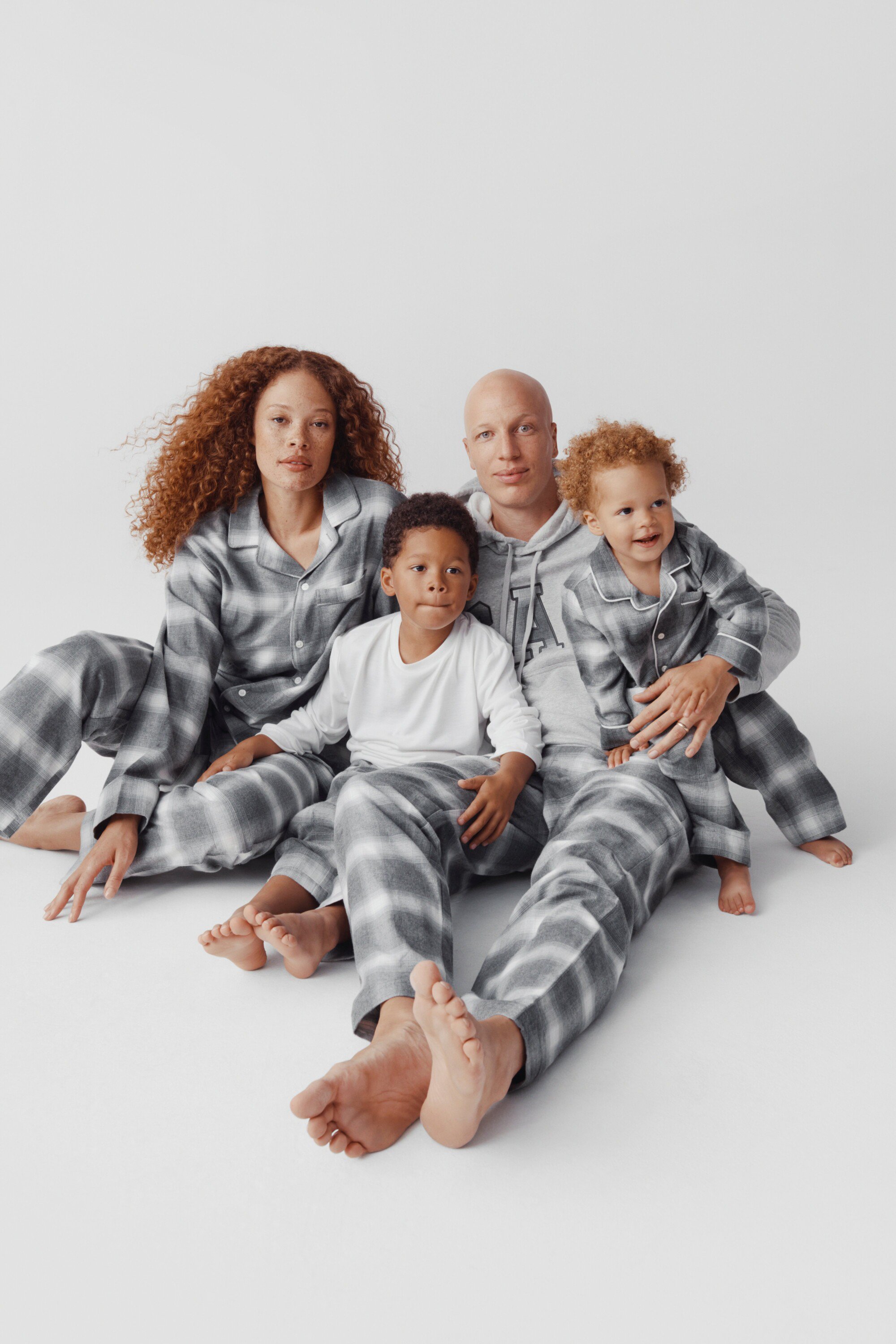Family of four poses in matching grey patterned pajamas against a white backdrop. Two adults and two children, indoor setting, pajamas.