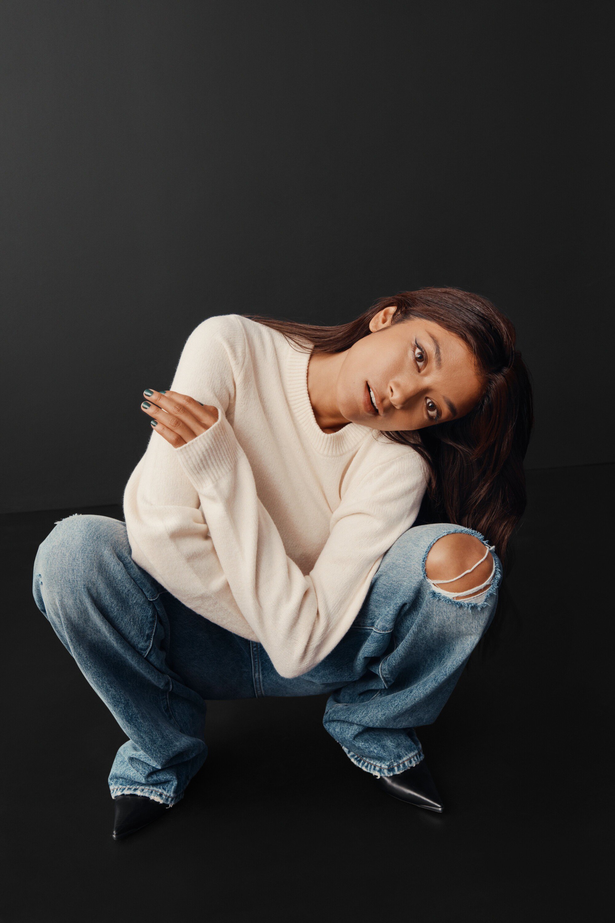 A young adult woman with long brown hair is crouching with her arms crossed, wearing a cream sweater and distressed denim jeans in a studio.