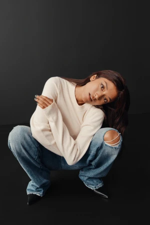 A young adult woman with long brown hair is crouching with her arms crossed, wearing a cream sweater and distressed denim jeans in a studio.