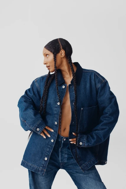Woman in denim jacket and jeans strikes a pose in a studio, looking left, with hands on hips and braids in her hair.