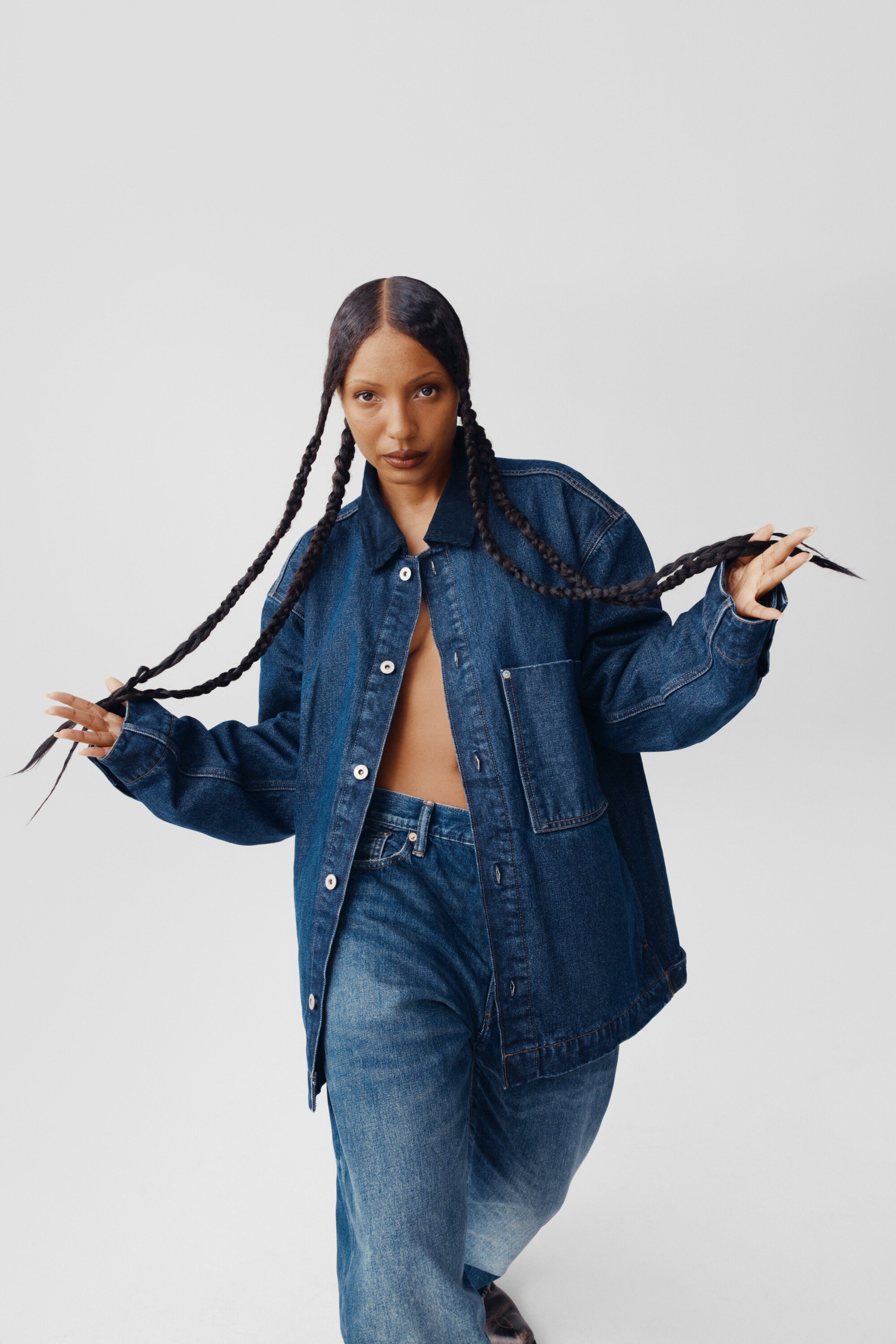 A woman with long braids poses in a blue denim jacket and jeans against a light background in a full shot, capturing a modern fashion vibe.