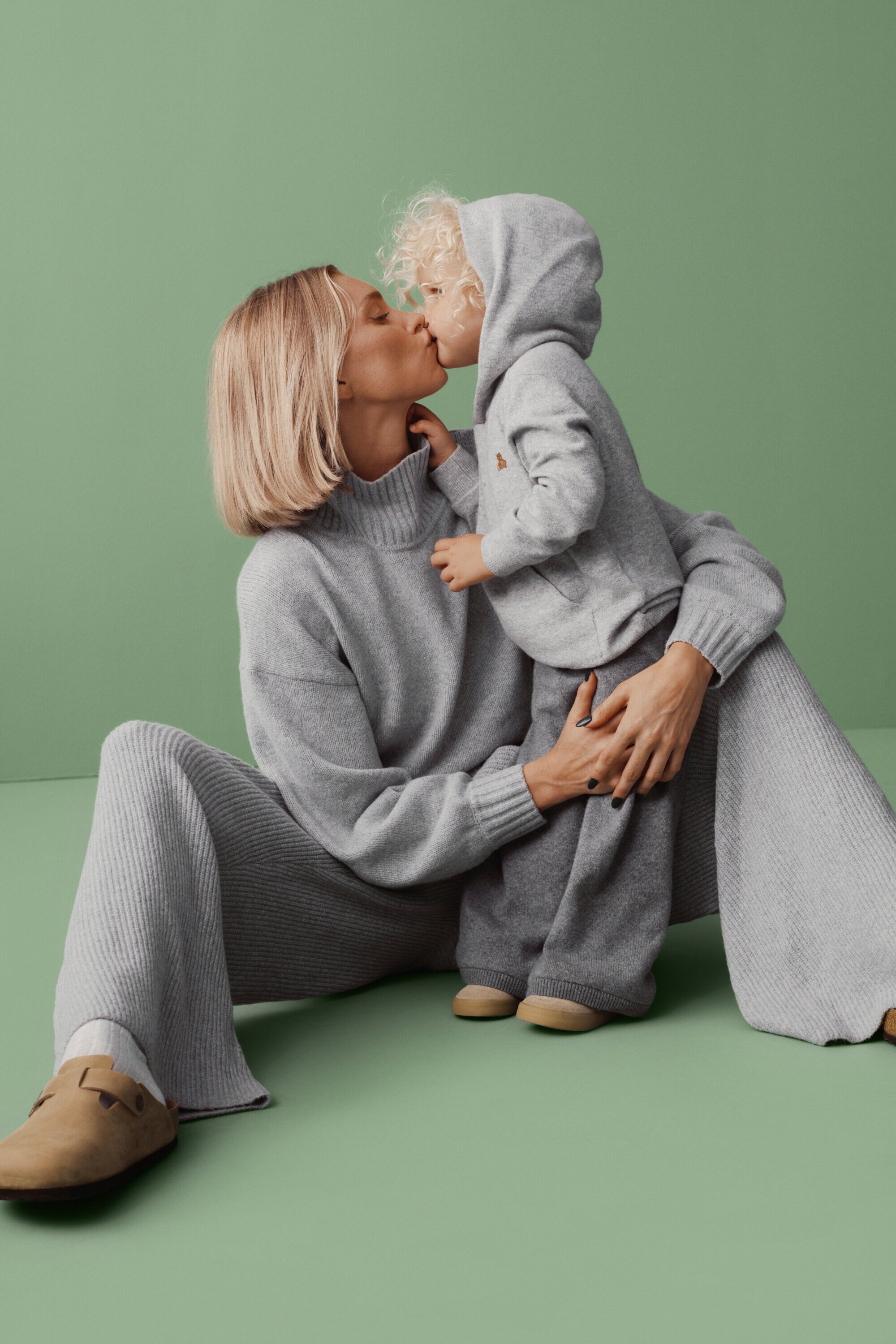 A blonde woman and child in matching grey knitwear share a kiss against a pastel green backdrop in a cozy, loving, indoor studio setting.