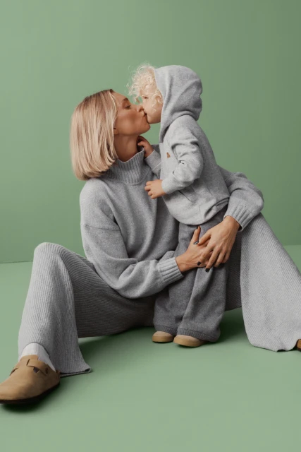 A blonde woman and child in matching grey knitwear share a kiss against a pastel green backdrop in a cozy, loving, indoor studio setting.