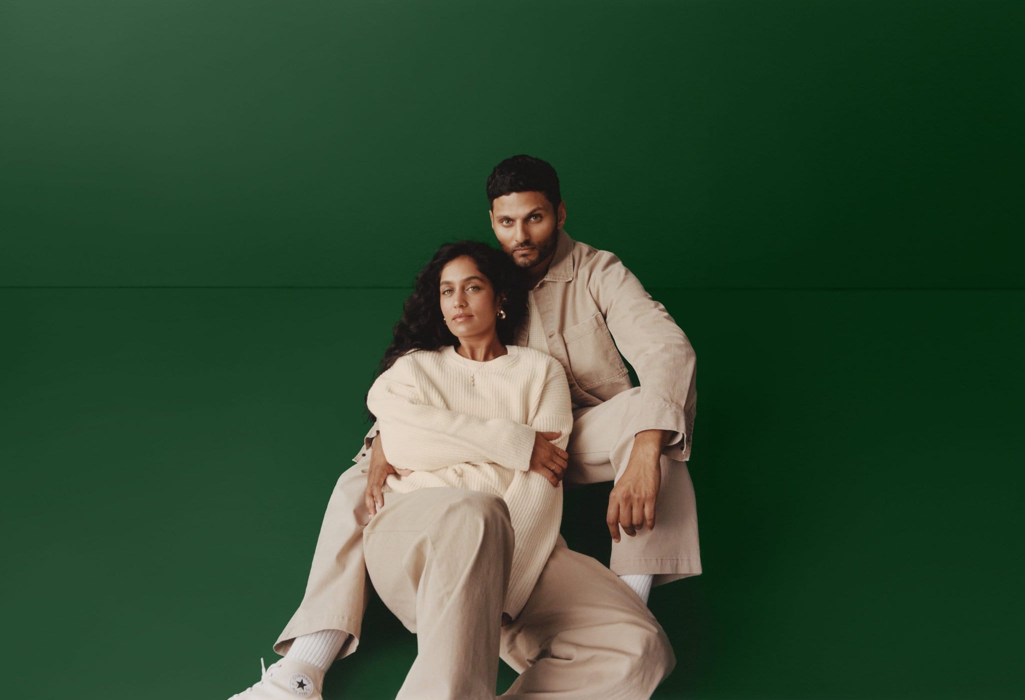 Jay Shetty and Radhi Devlukia seated together wearing neutral beige and cream Gap clothing against a deep green backdrop.