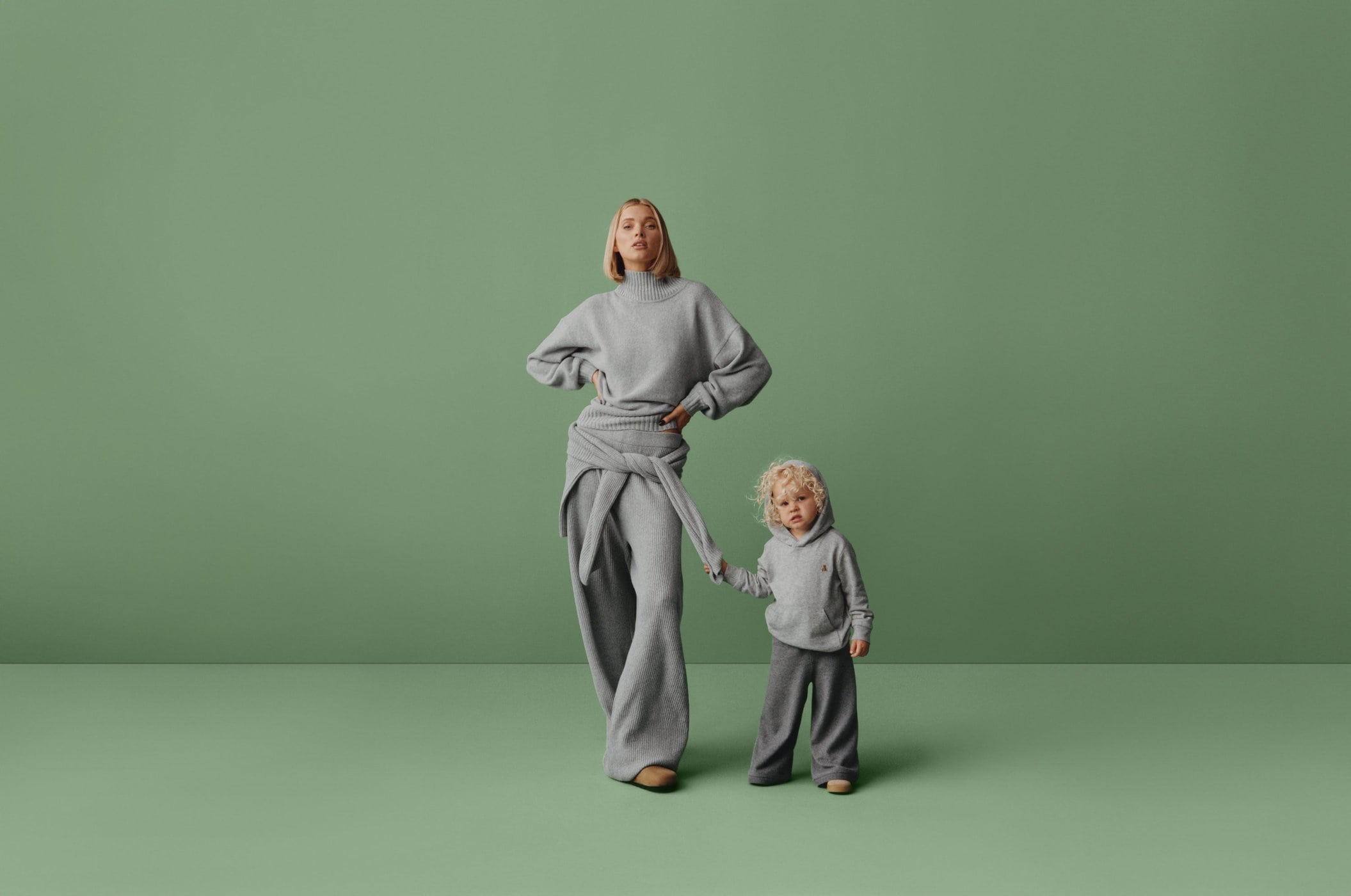 A woman and a child stand side by side in a studio, both dressed in matching grey knitwear against a solid green backdrop.