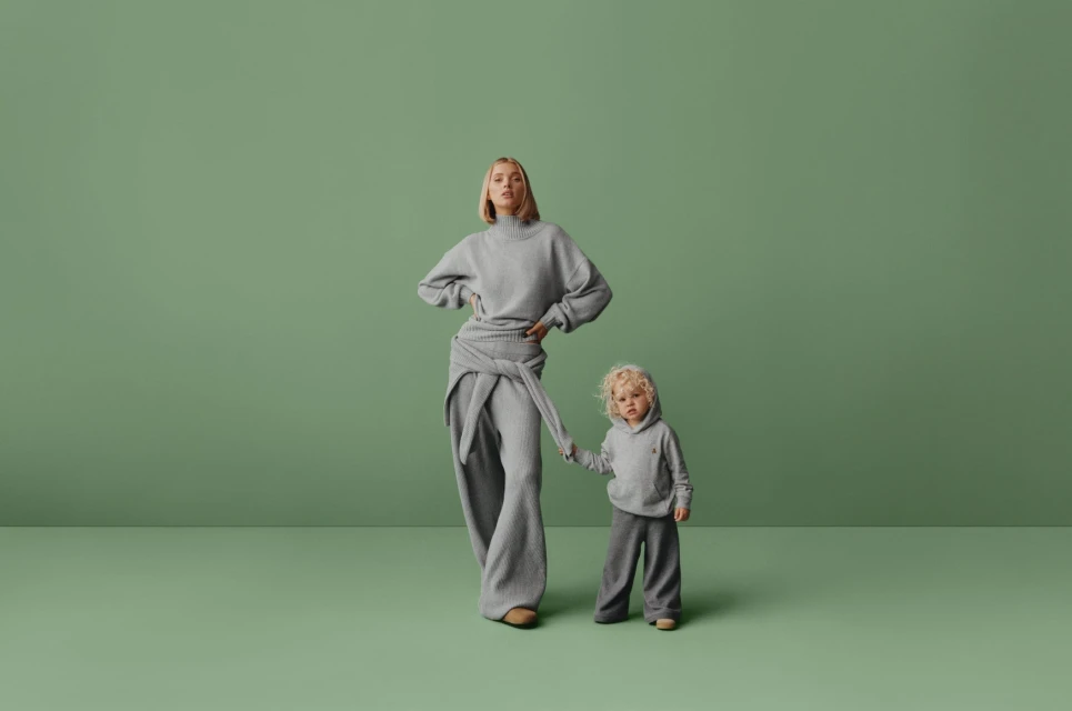 A woman and a child stand side by side in a studio, both dressed in matching grey knitwear against a solid green backdrop.