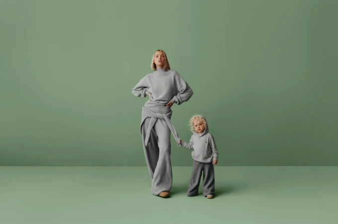 A woman and a child stand side by side in a studio, both dressed in matching grey knitwear against a solid green backdrop.