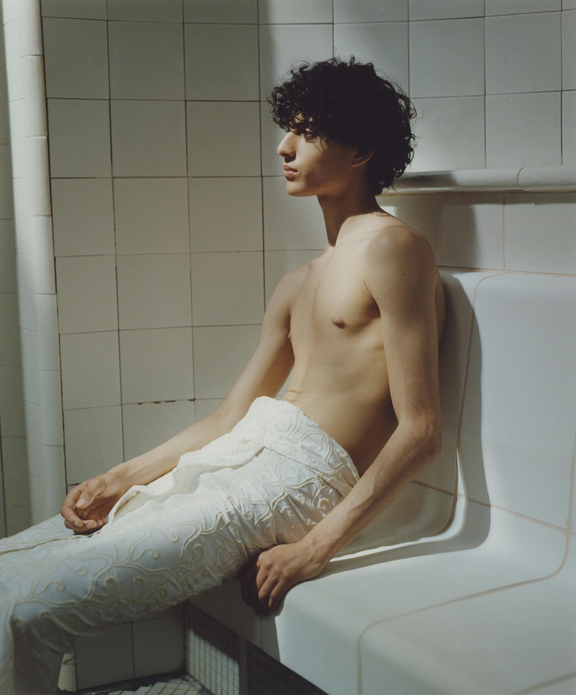 Young adult male with curly hair sits shirtless on a white bench in a tiled room with natural light streaming from the left.