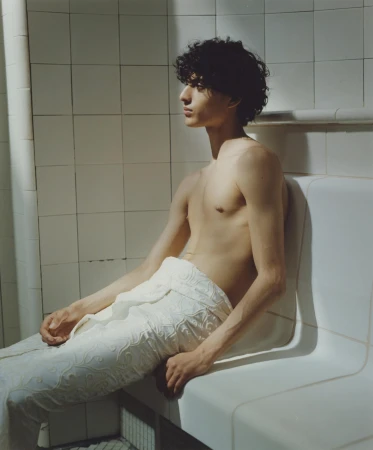 Young adult male with curly hair sits shirtless on a white bench in a tiled room with natural light streaming from the left.
