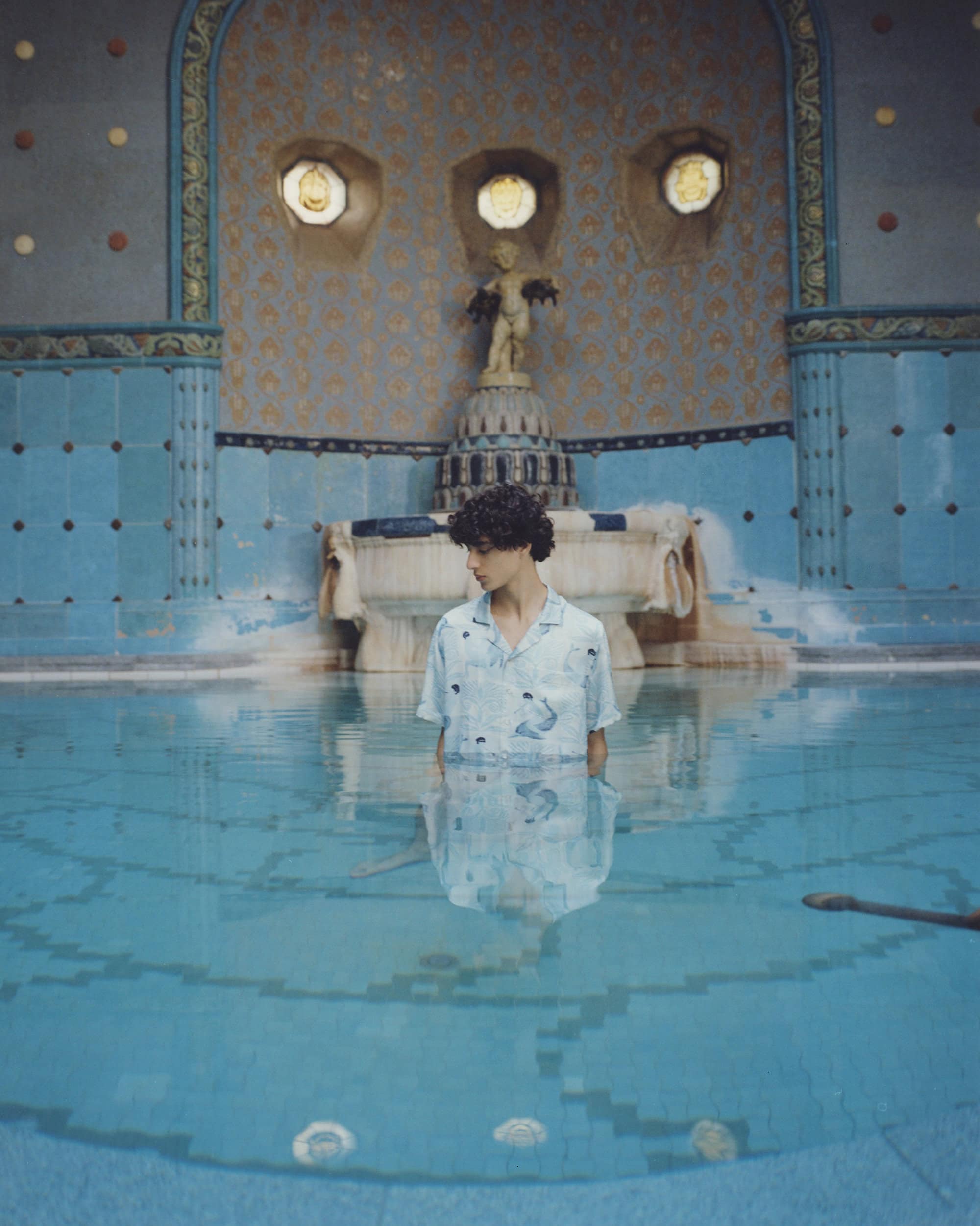 Man standing waist-deep in a bright blue pool with patterned tile walls, statue, and decorative wall features in the background.