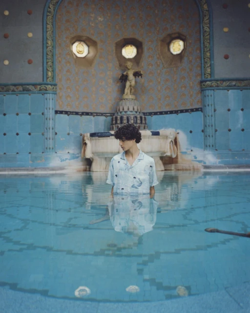 Man standing waist-deep in a bright blue pool with patterned tile walls, statue, and decorative wall features in the background.