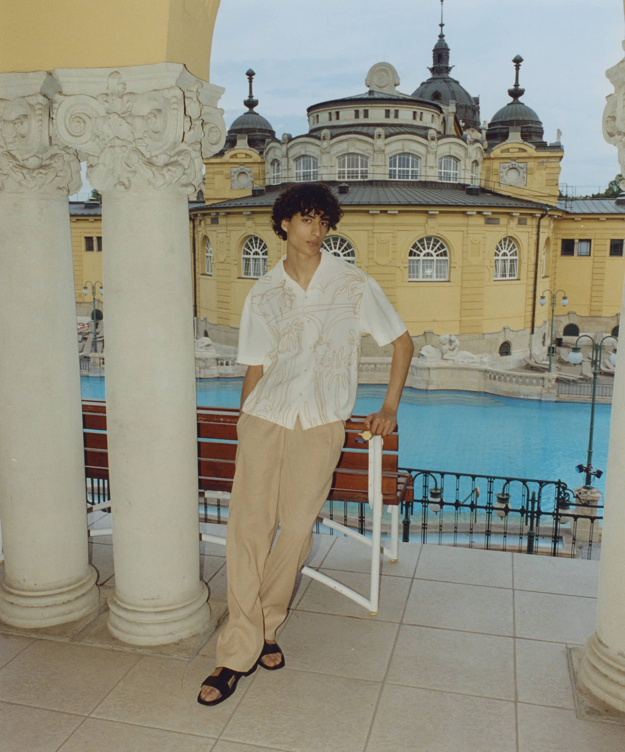 Young man poses in patterned shirt and tan pants on a terrace with columns, overlooking an outdoor pool and ornate building in Budapest.