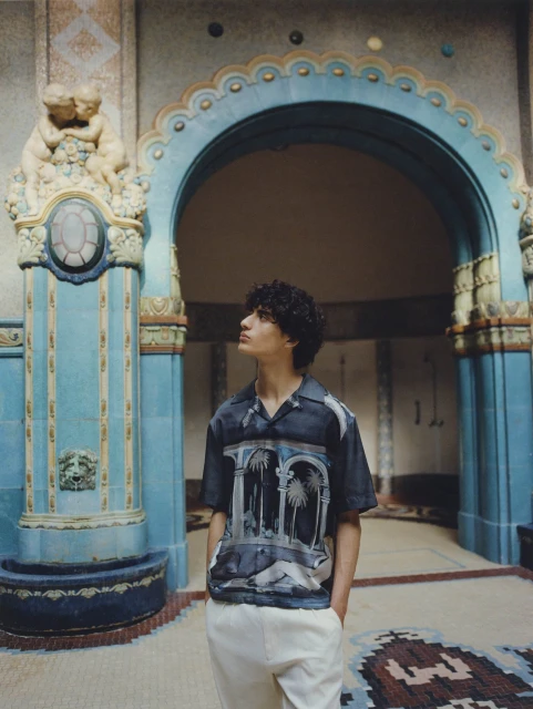 Young man in printed short-sleeve shirt with cream pants stands in an ornate hall with columns, archways, and tiled floors.