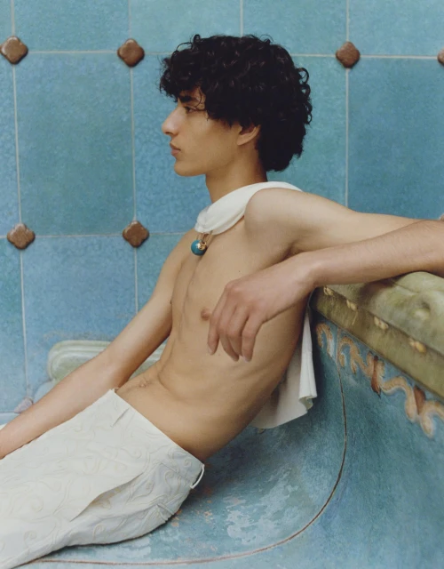 Young adult with curly hair and wearing white pants leans against a blue-tiled wall indoors, accessorized with a turquoise pendant necklace.