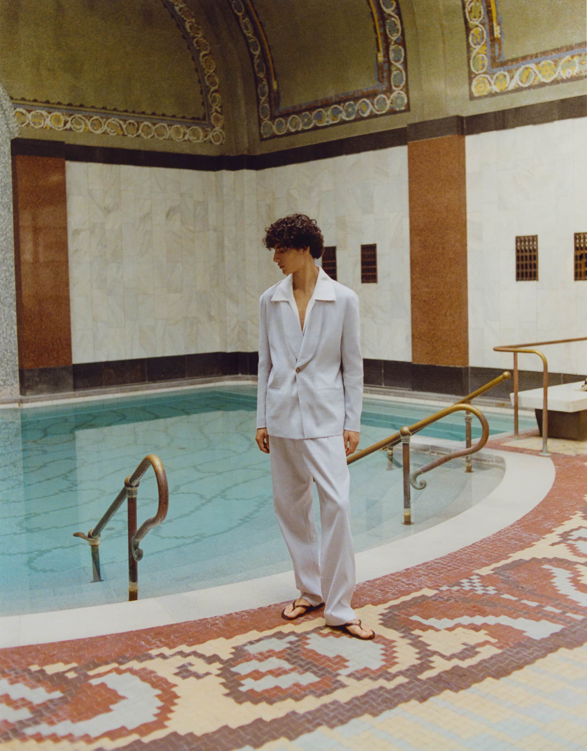 An adult male with curly hair is standing in a light-colored suit next to a pool inside an ornate tiled building with sunlight filtering through.