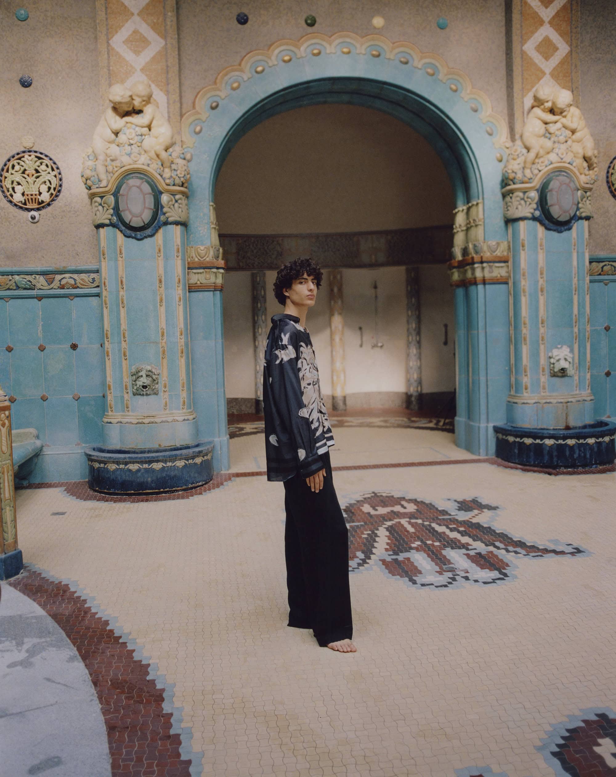 Man in floral top poses in ornate building with archways and patterned tile floor, creating a vintage fashion editorial look.