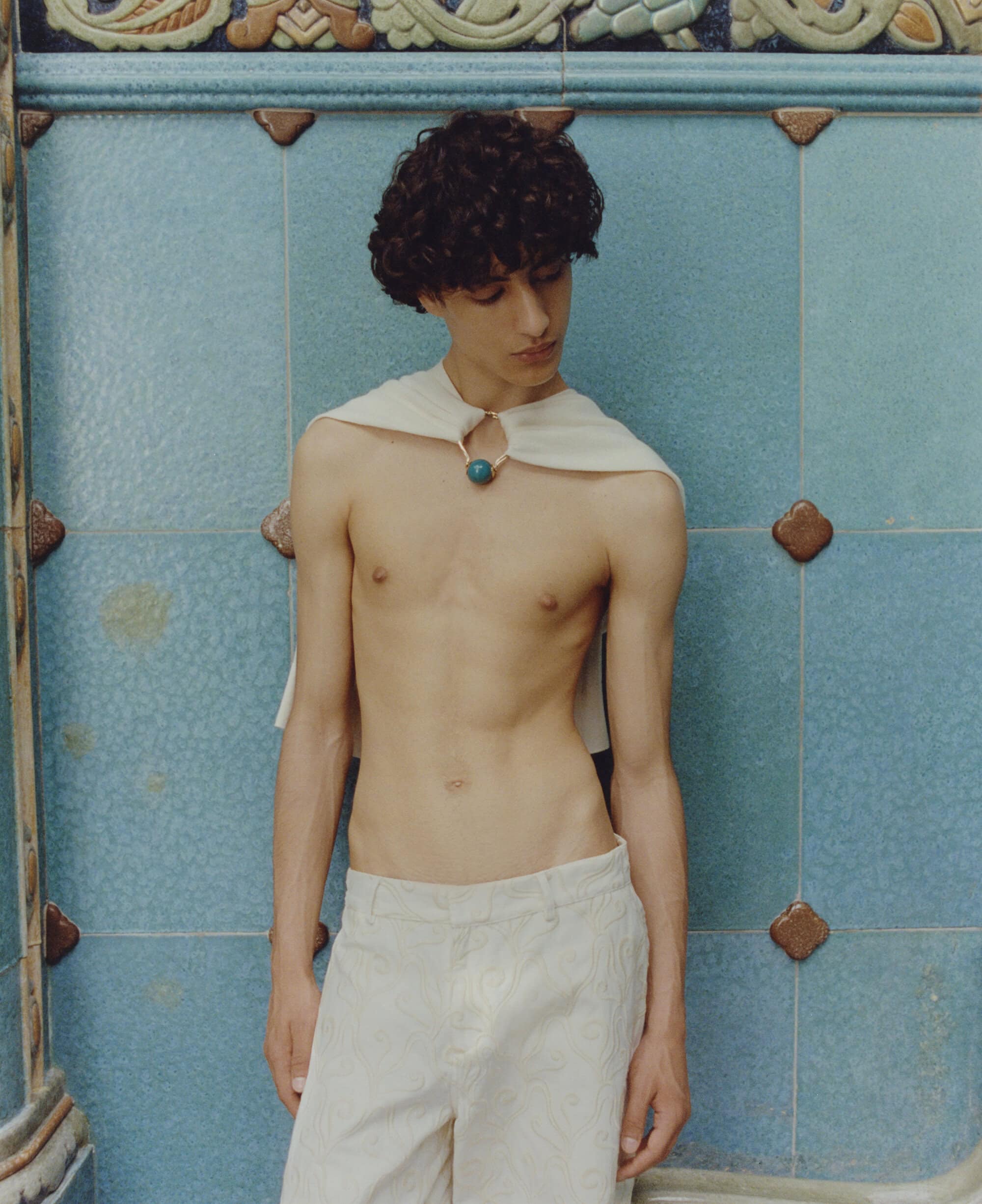 Young adult poses shirtless in white trousers and draped fabric against a blue tiled wall with geometric details in a fashion-editorial shot.