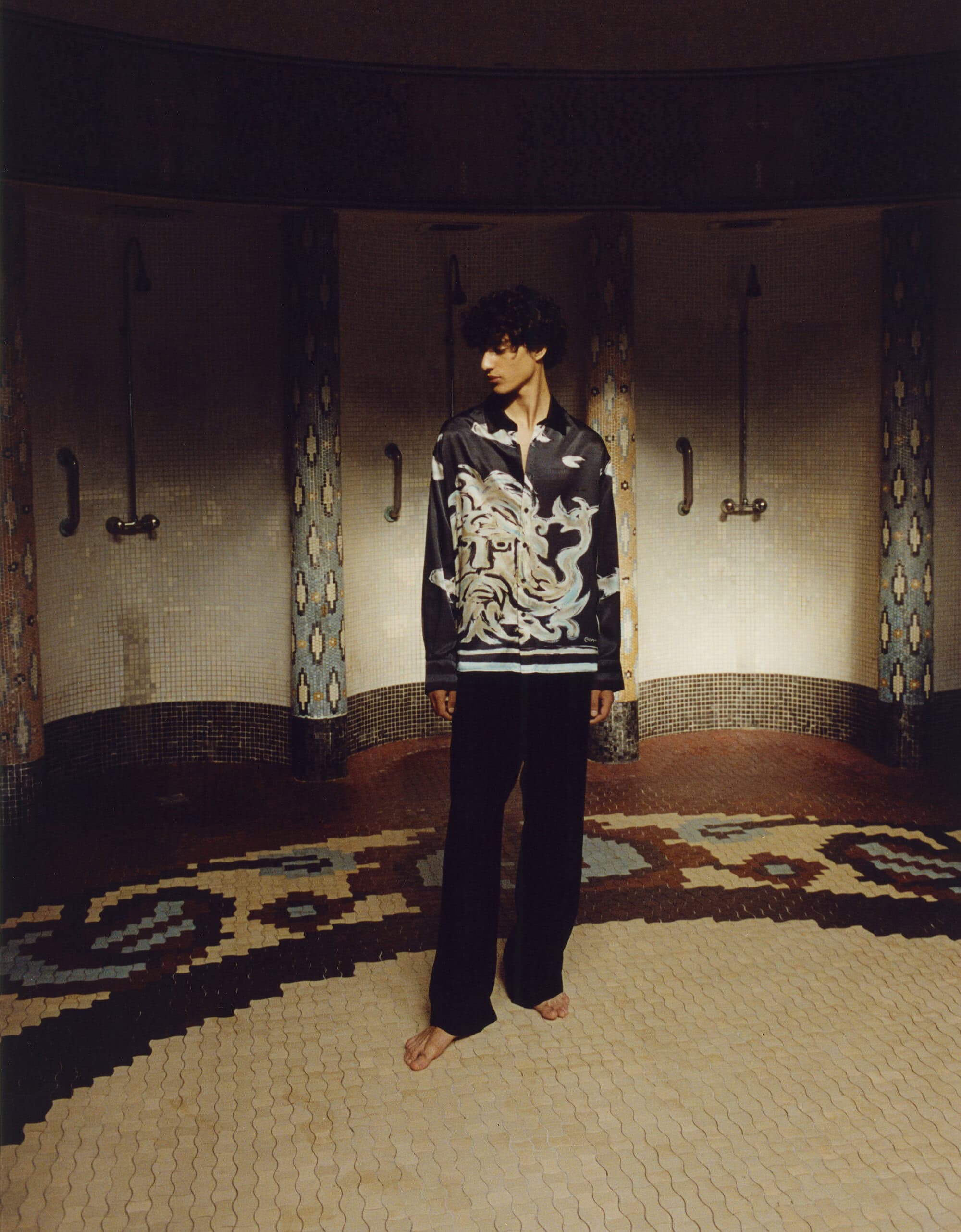 Young adult stands barefoot in vintage-tiled bathhouse wearing pajamas-style graphic shirt and pants in a dimly lit indoor space.