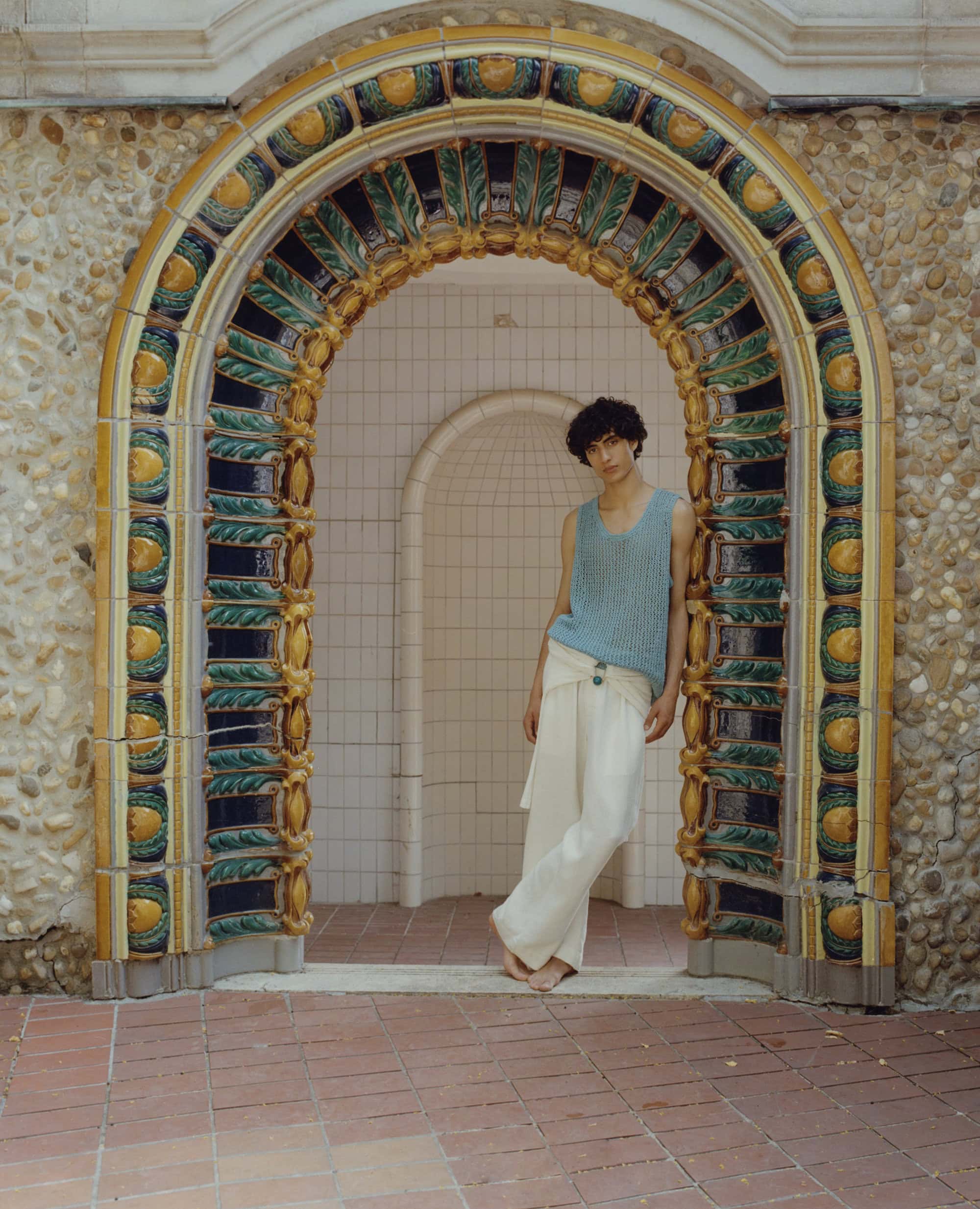 Young adult stands in archway with decorative tile work wearing light blue knit top and white linen pants, outdoor shot, eye-level angle.
