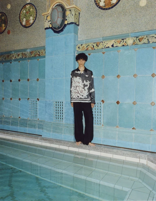 Young adult in patterned blouse and black pants stands barefoot beside tiled indoor pool with decorative tile and turquoise water.
