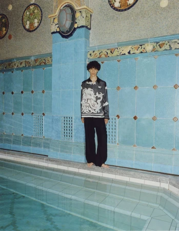 Young adult in patterned blouse and black pants stands barefoot beside tiled indoor pool with decorative tile and turquoise water.
