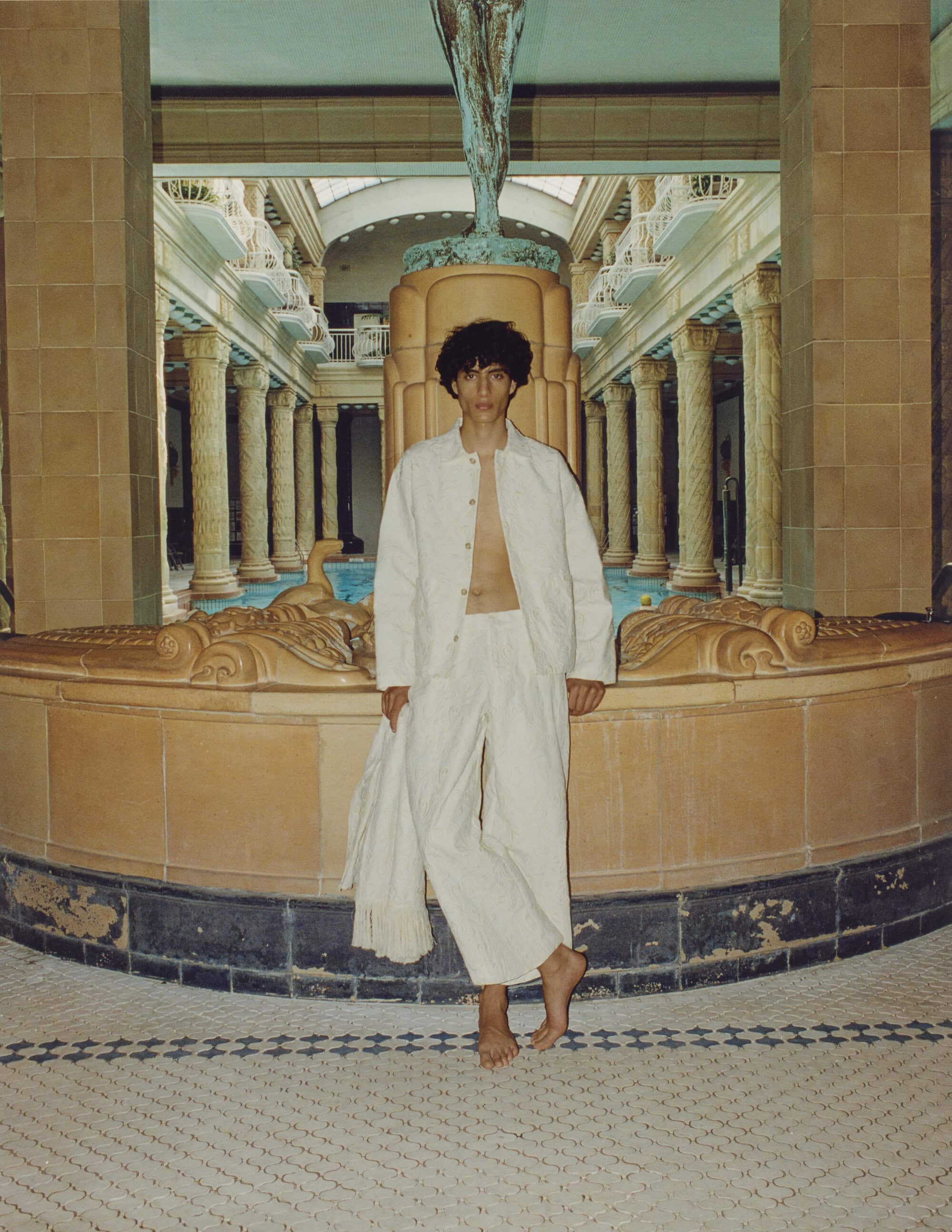Young man in white linen suit stands barefoot near a large indoor fountain with classical columns and tiled floor in neutral colors.