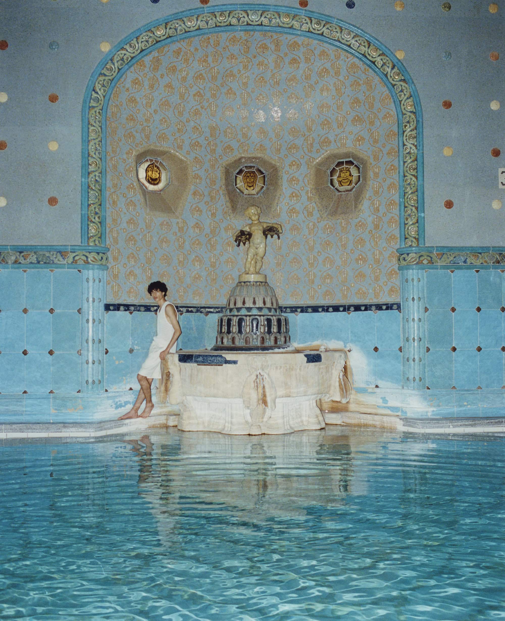 Young adult sits on the edge of a turquoise tiled pool with decorative trim and a small statue in an indoor pool environment.