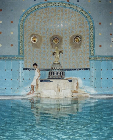 Young adult sits on the edge of a turquoise tiled pool with decorative trim and a small statue in an indoor pool environment.