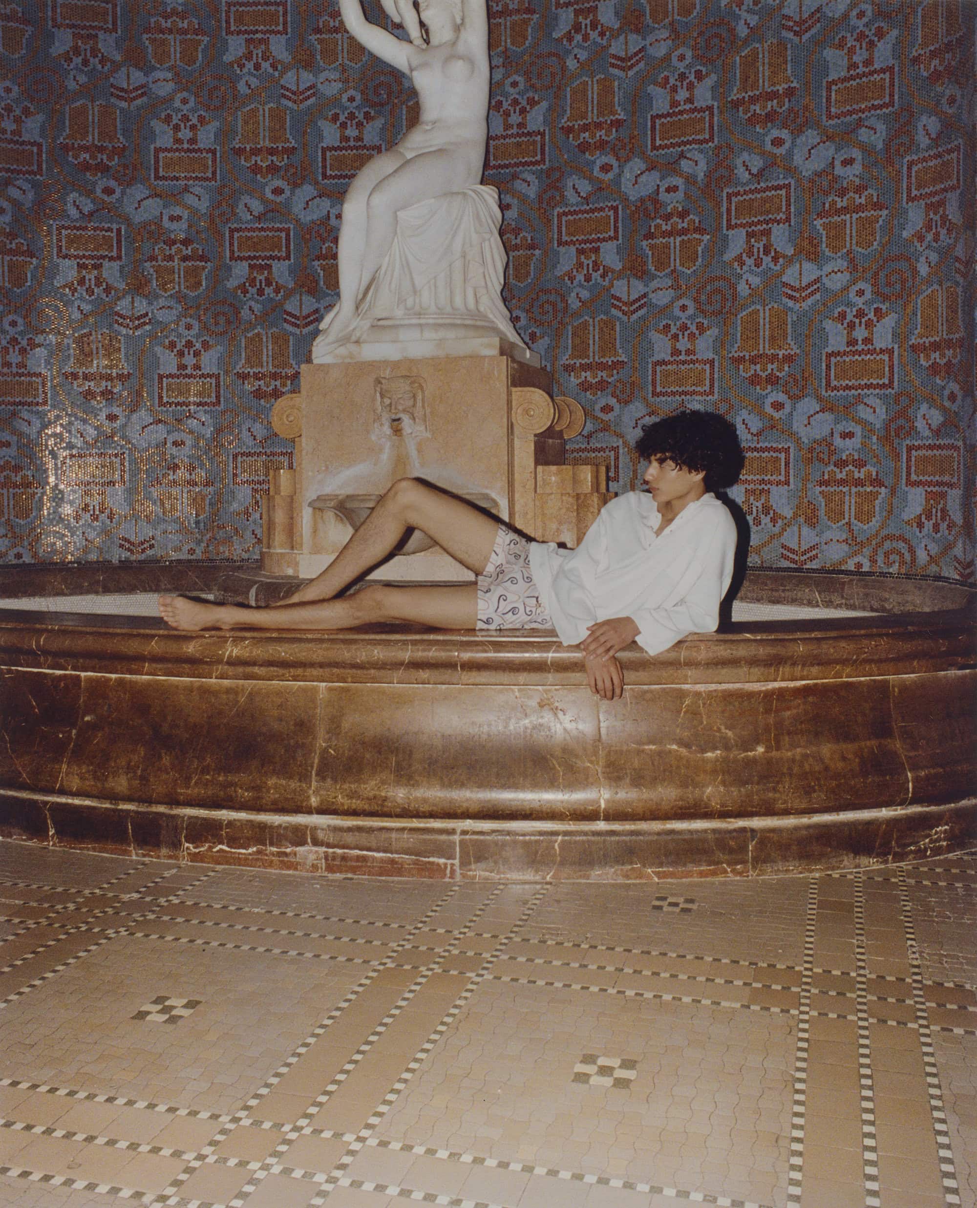 An adult lies in a marble fountain in an indoor setting, posed beside a white statue against a backdrop of patterned tiles.