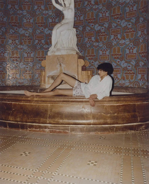 An adult lies in a marble fountain in an indoor setting, posed beside a white statue against a backdrop of patterned tiles.