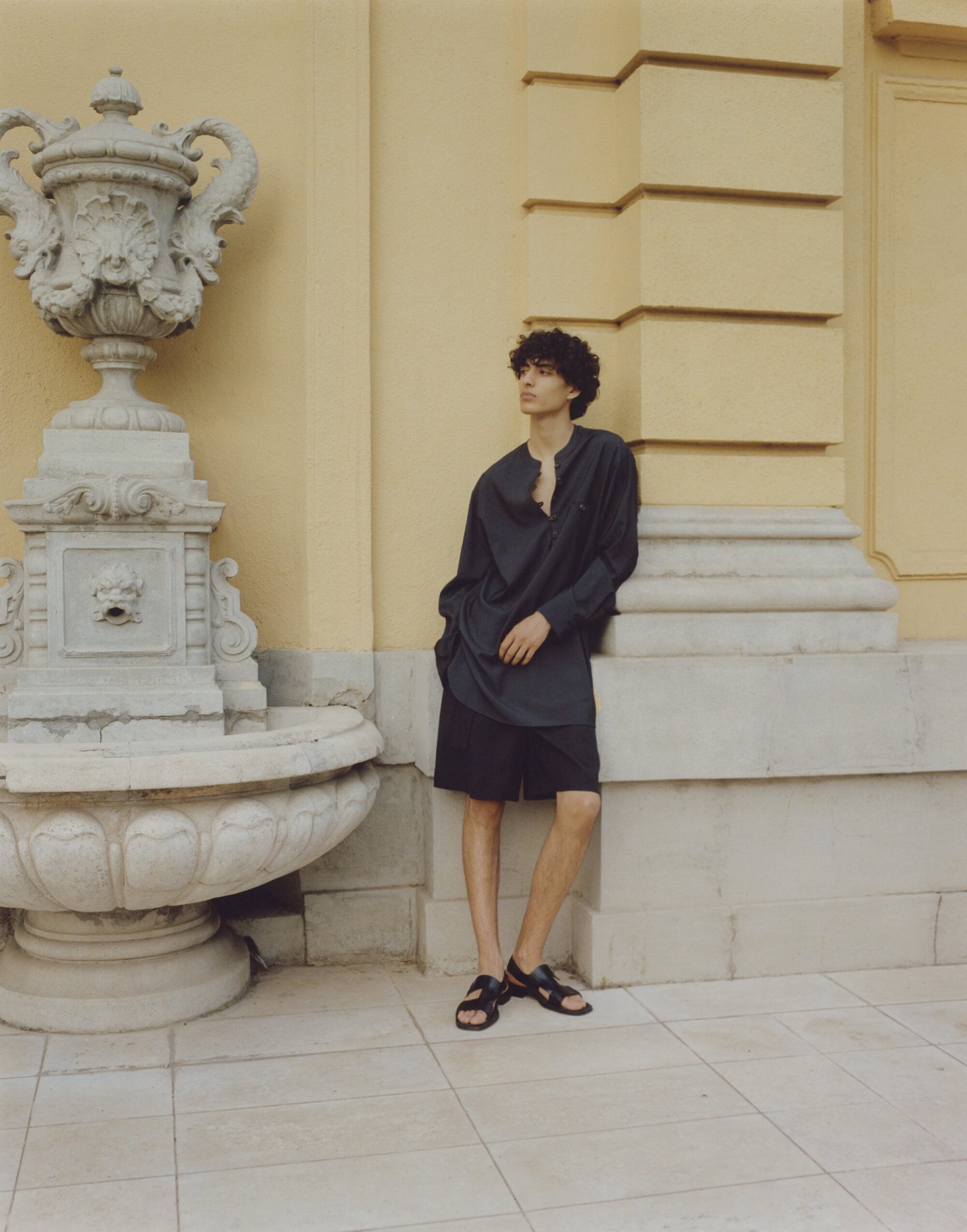 A young man leans against a yellow wall near a decorative fountain. He wears a black tunic, black shorts, and black sandals.