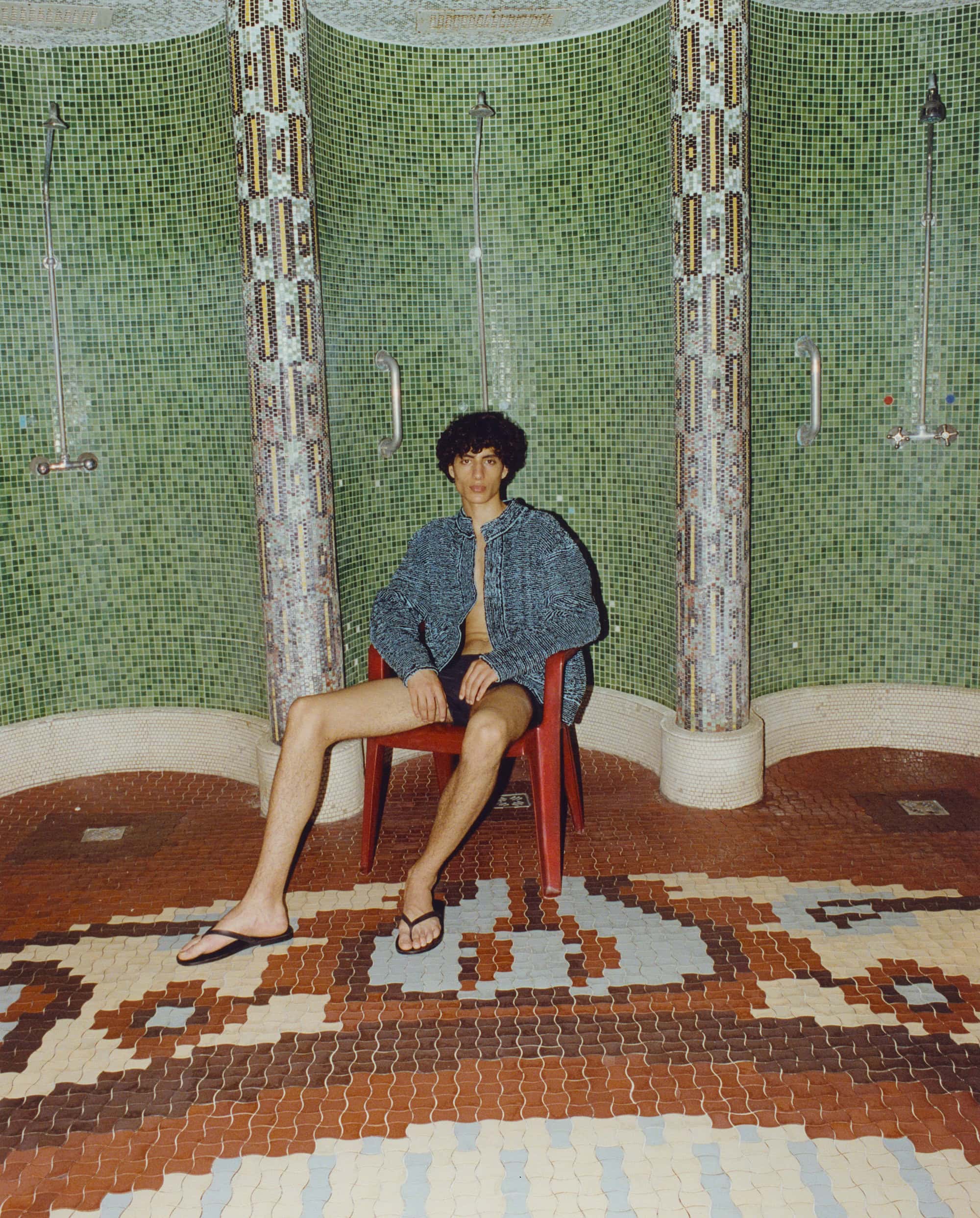 Young adult male with dark curly hair sits on a red chair in a tiled shower room wearing a blue patterned shirt and black flip-flops.
