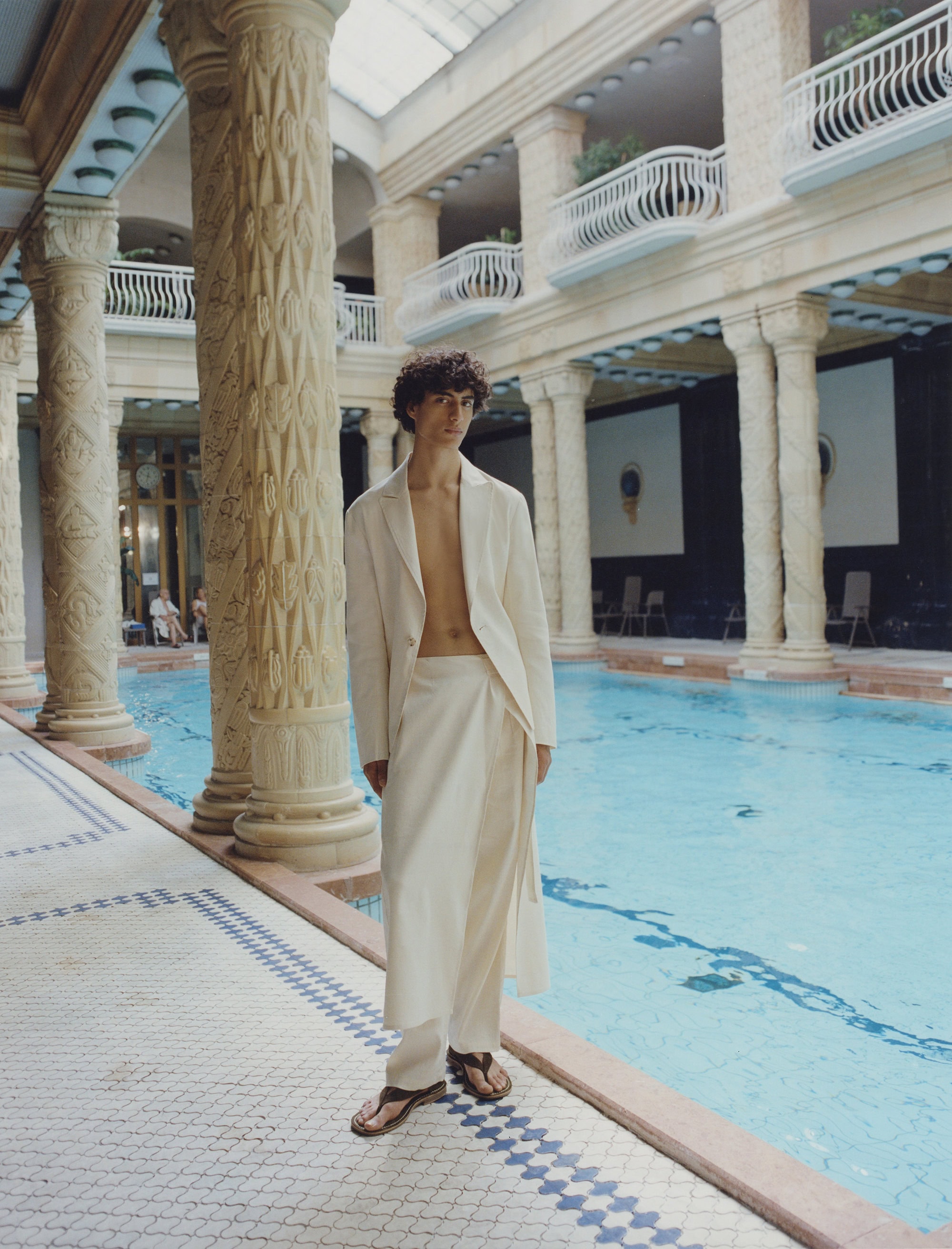 Adult male in cream-colored linen coat and wrap skirt poses by pool indoors with overhead lighting and tiled floor and column details.