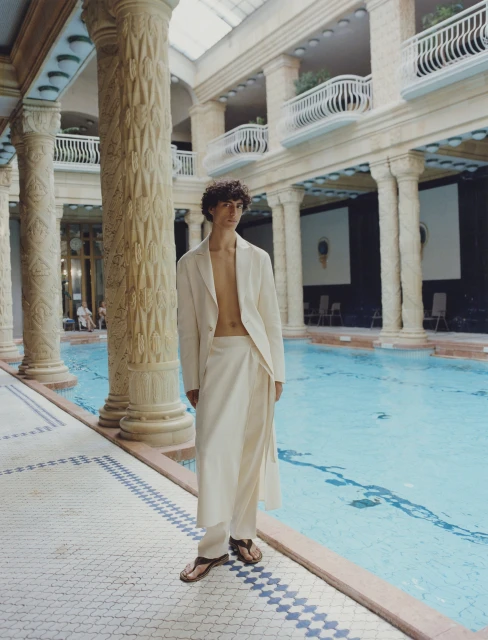 Adult male in cream-colored linen coat and wrap skirt poses by pool indoors with overhead lighting and tiled floor and column details.