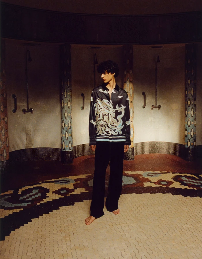 Model standing barefoot in a patterned tiled room wearing a black printed silk shirt and dark trousers for COMMAS Resort 2024