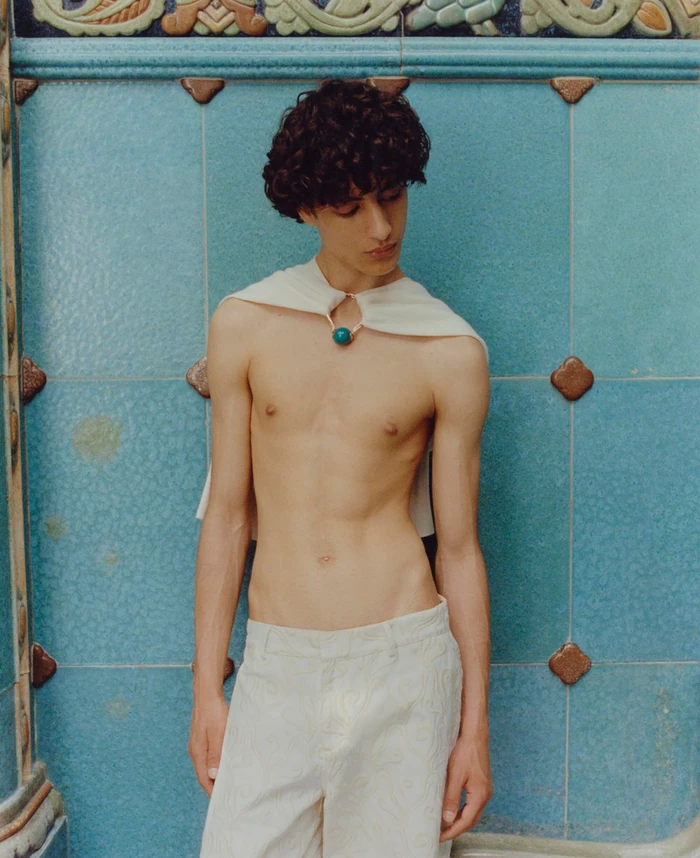 Model Yoesry Detre standing shirtless in off-white trousers and a white capelet against a turquoise tiled wall for COMMAS Resort 2024.